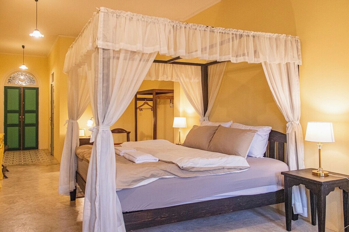 Uma cama com dossel e cortinas, roupa de cama branca, uma mesa de cabeceira com abajur e uma porta verde ao fundo.