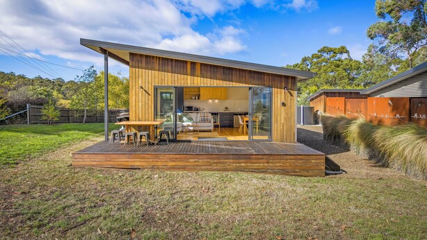 Una casa moderna con terraza de madera, cocina con puertas corredizas de vidrio y patio trasero con césped y árboles.