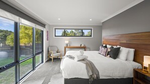 Habitación de hotel moderna con una cama grande, un cabecero de madera, una mesita de noche, una silla y una ventana con vistas a árboles.