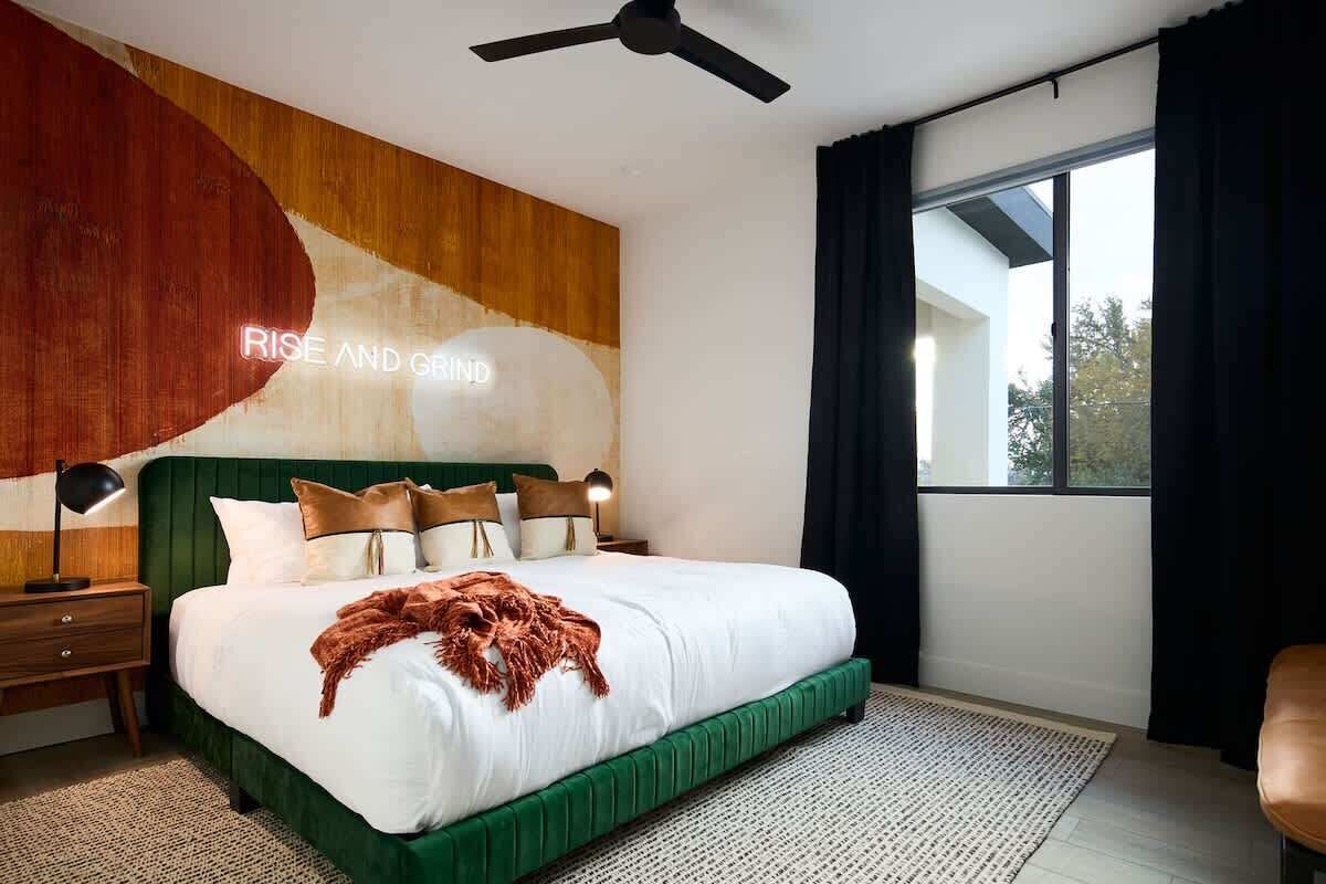 A bedroom with a bed, a ceiling fan, a nightstand, a lamp, a window with curtains, and a wall with a large abstract painting.