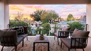 Ein Balkon mit einer Couch, Stühlen und einem Tisch mit Getränken, mit Blick auf ein Wohngebiet mit Bäumen und Gebäuden.