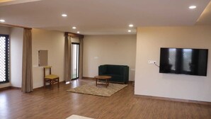 A spacious living room with a flat-screen TV mounted on the wall, a green sofa, a wooden coffee table, and a yellow chair. - Devraha Seva Ashram - Vrindavan (Mathura)