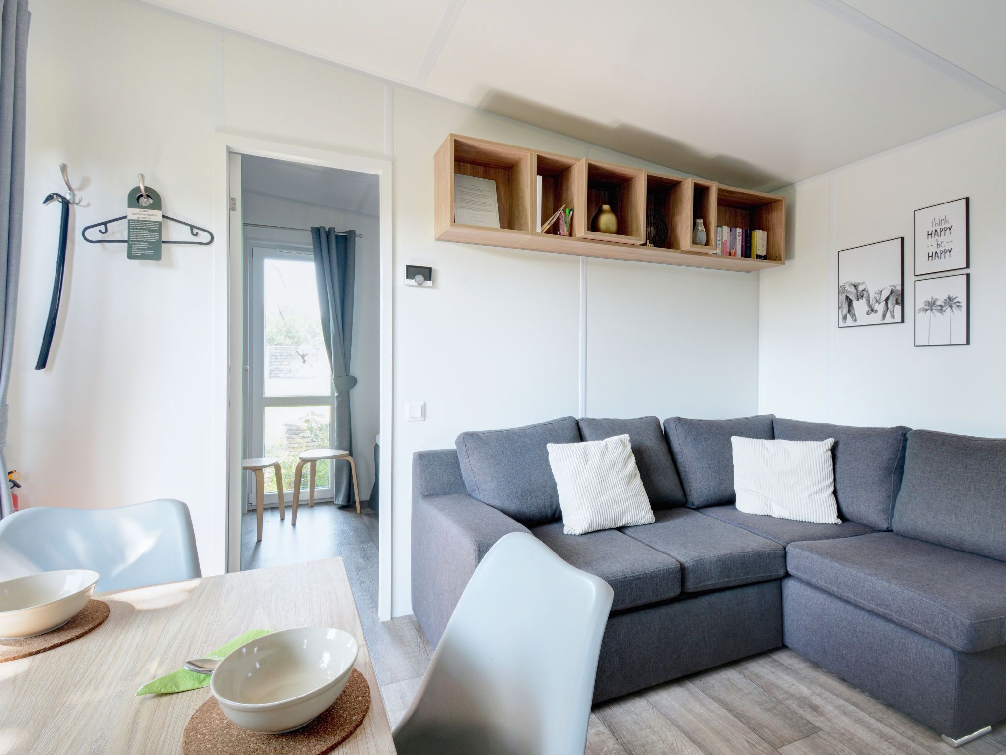 A modern living room with a grey sofa, two white pillows, a wooden shelf with books and decorative items, and a dining area with a table and chairs.