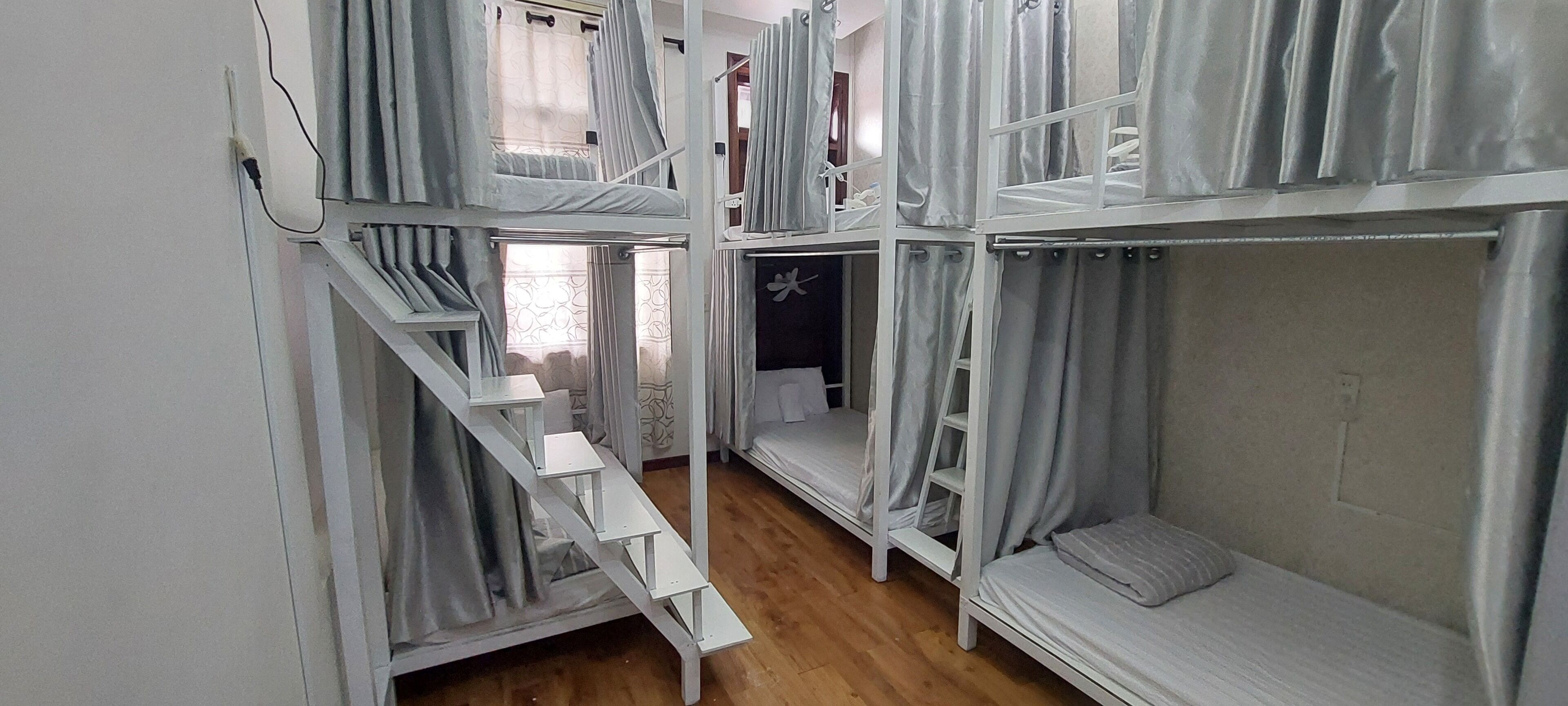 A dormitory-style room with bunk beds, a staircase, and a small window with curtains.