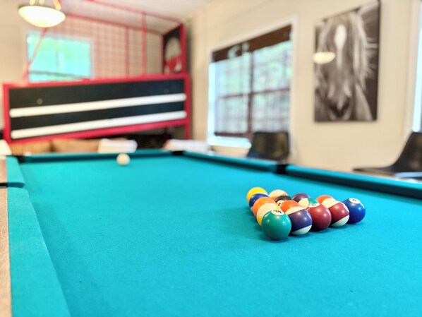 A pool table with colorful balls in a room with a red and black padded bench, a window with blinds, and a black and white artwork on the wall. - Big family friendly, Deck, Pool Table, Propane Grill & Fire Pit! (Hot Springs)