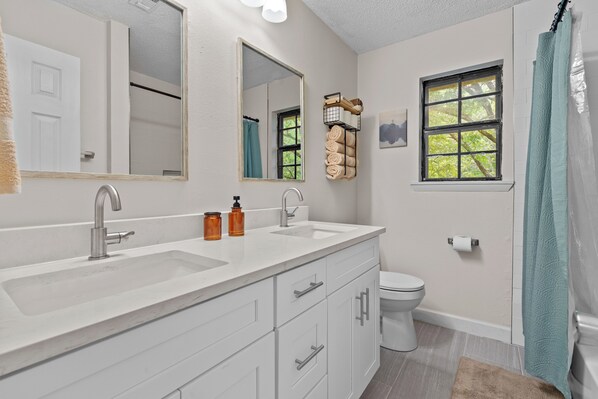 Une salle de bain avec un lavabo blanc, deux miroirs, des toilettes et une fenêtre donnant sur des arbres.