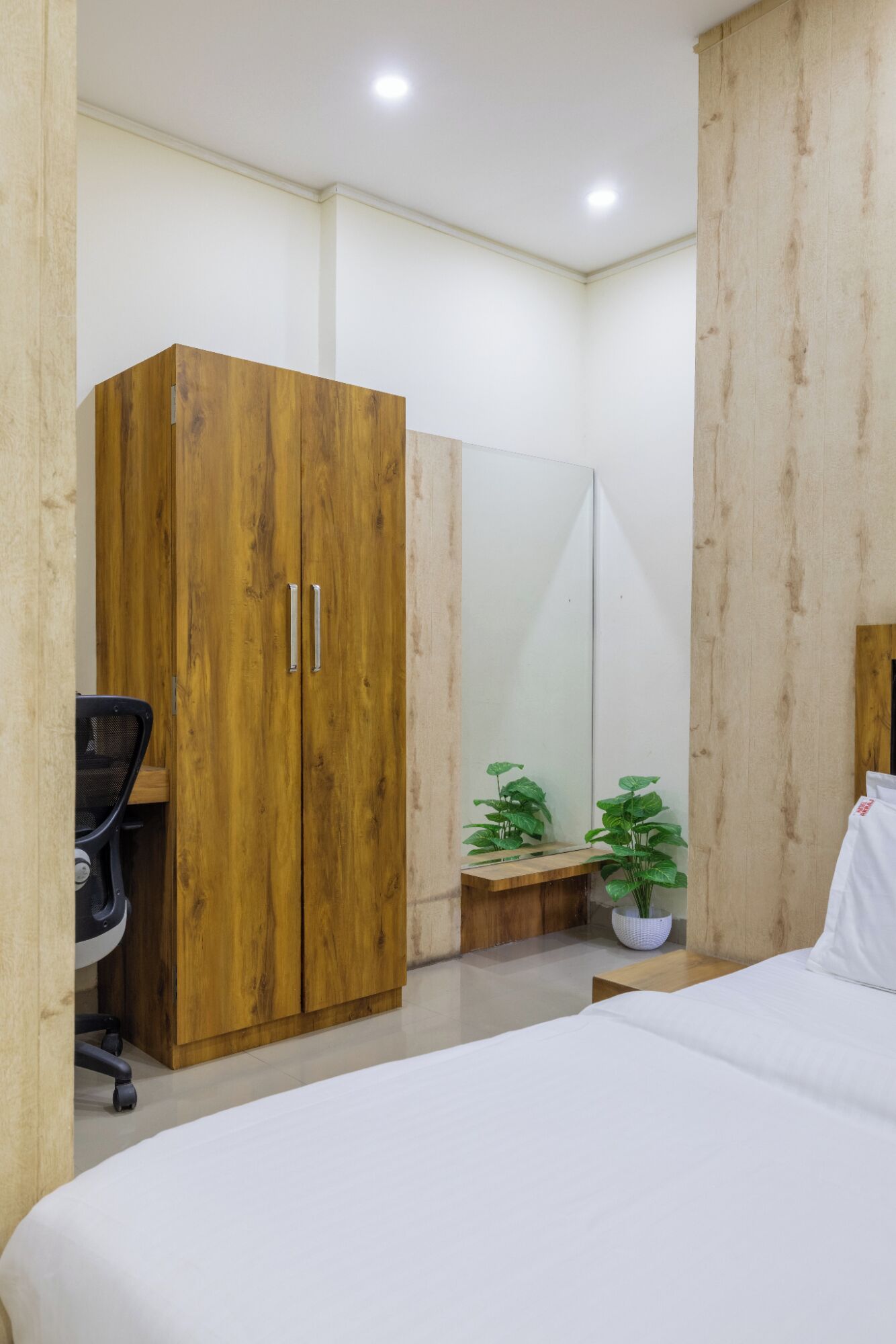 Un dormitorio con cama, armario de madera, escritorio con silla y dos plantas en macetas.