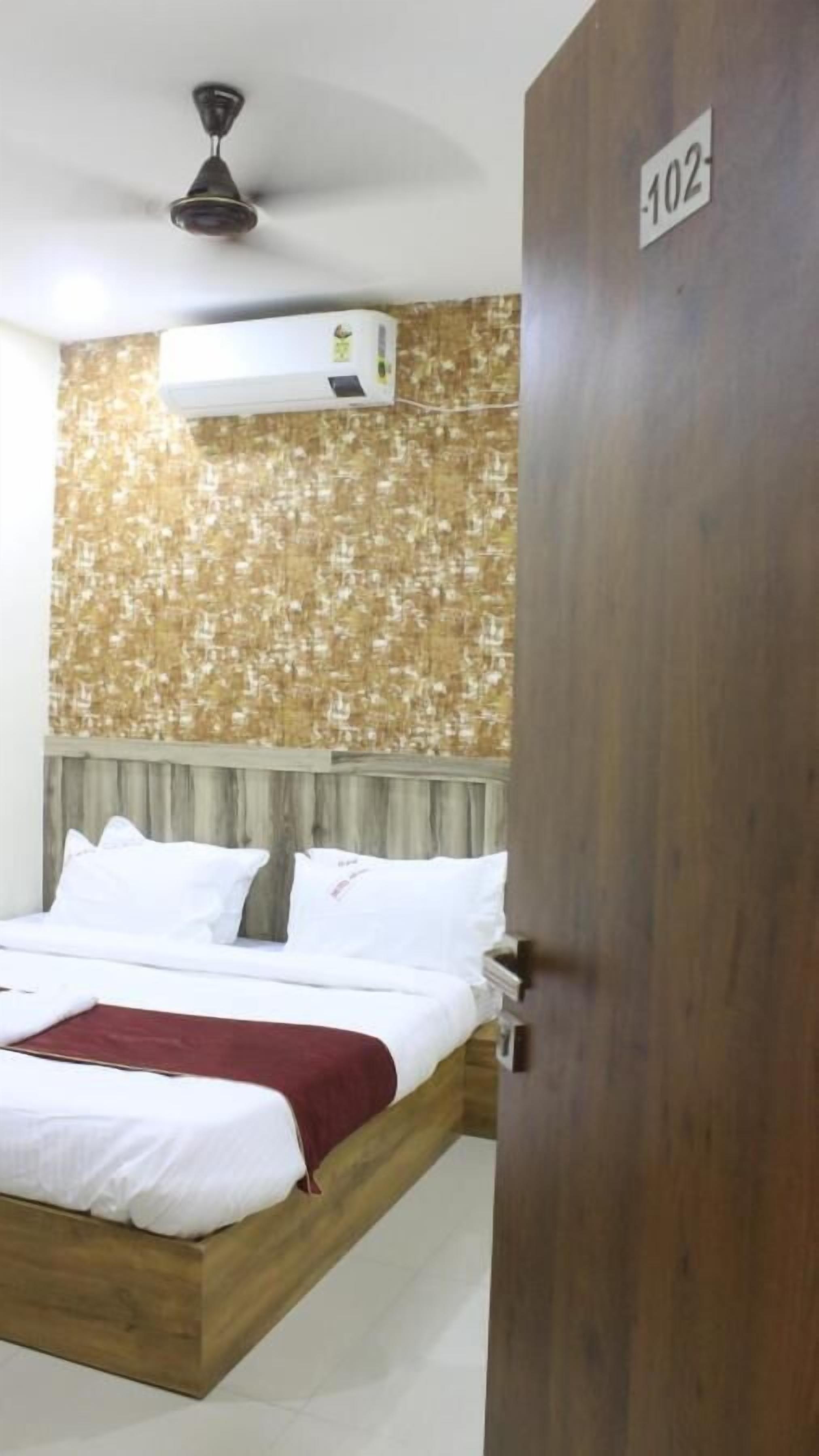 A hotel room with a bed, a ceiling fan, an air conditioning unit, and a wooden door with a number sign.