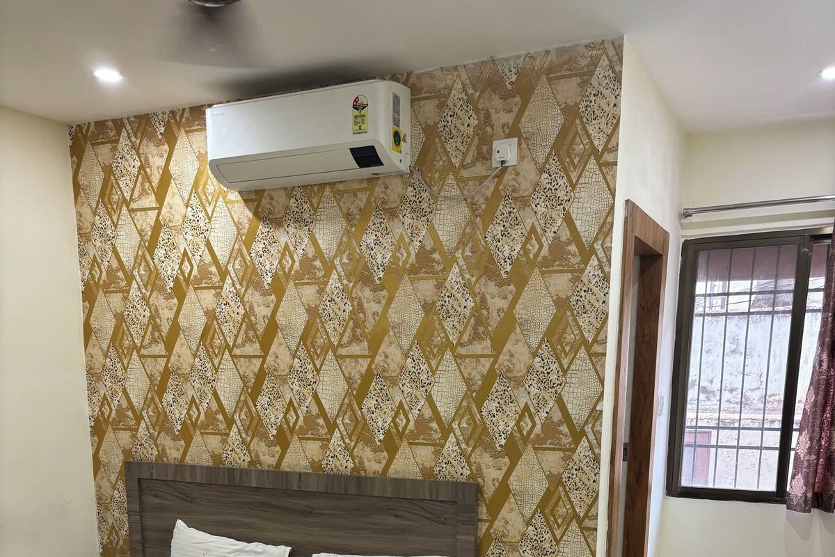 A bedroom with a bed, an air conditioner, a bedside table with a phone, and a window with a view.