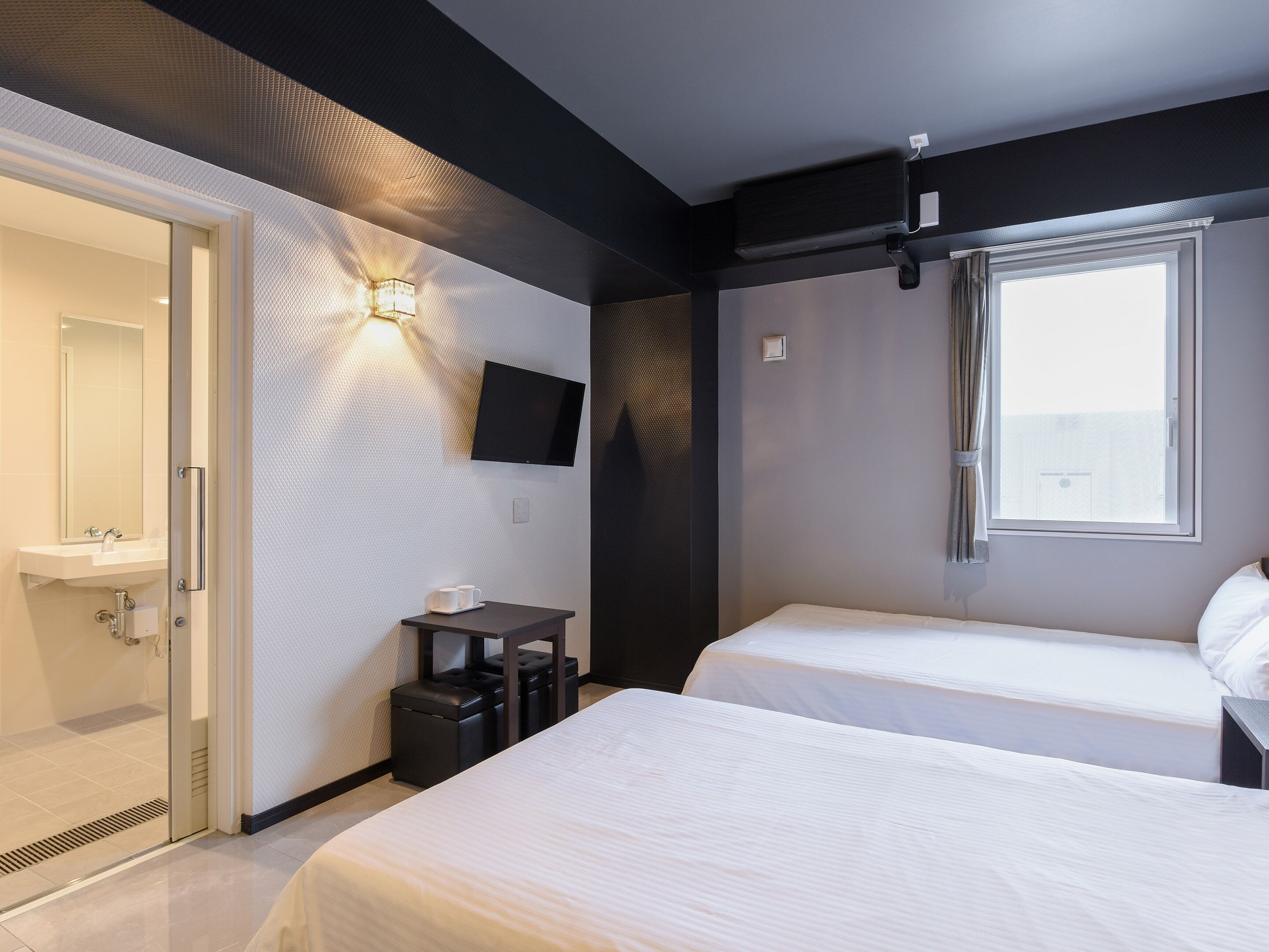 Quarto de hotel com duas camas, televisão na parede, uma mesinha com chávenas e uma janela com cortinas.