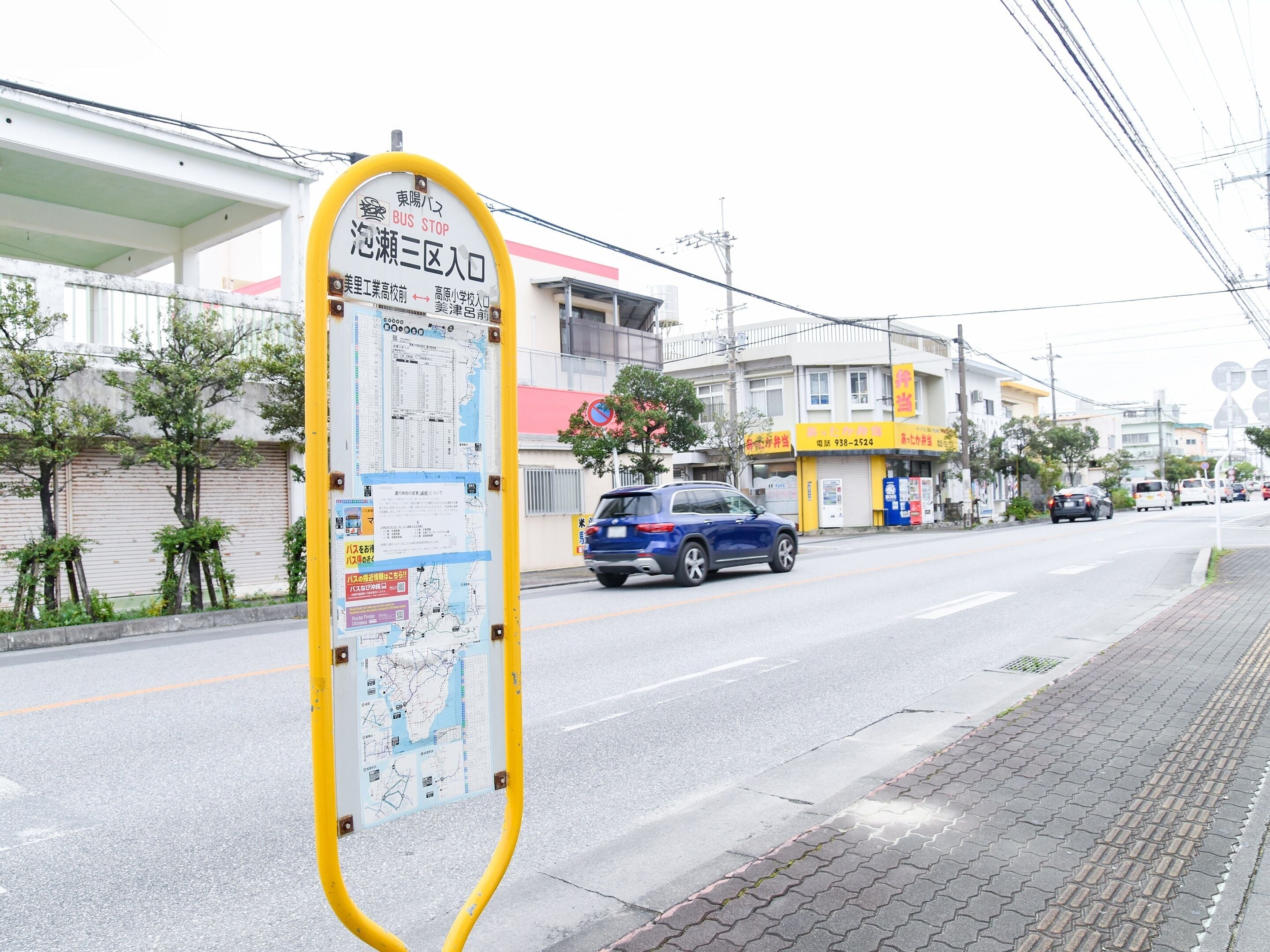 A bus stop sign with maps and directions, a blue car parked on the street, and a yellow storefront with a red awning.