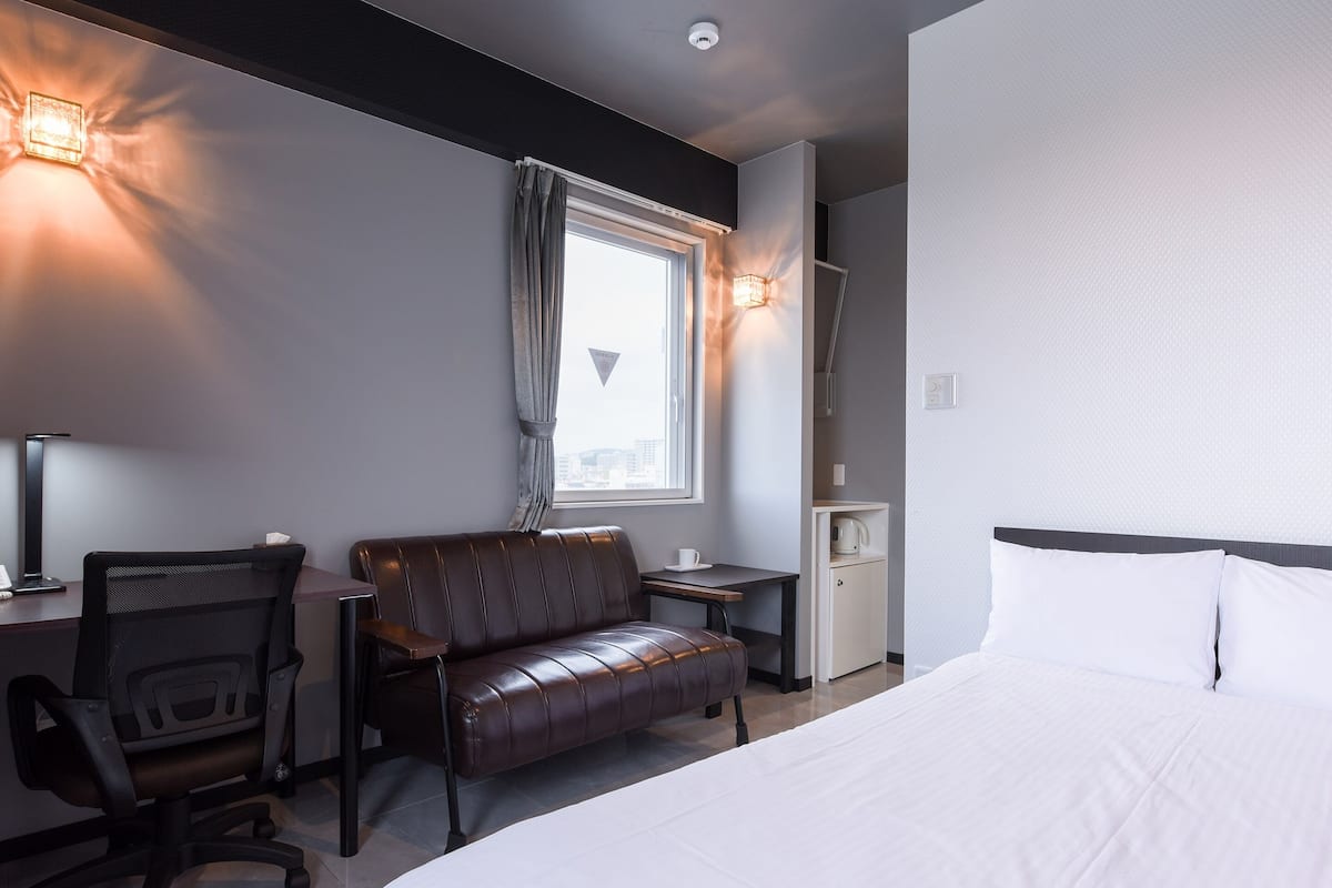 A hotel room with a bed, a desk with a chair, a small table, and a window with curtains.