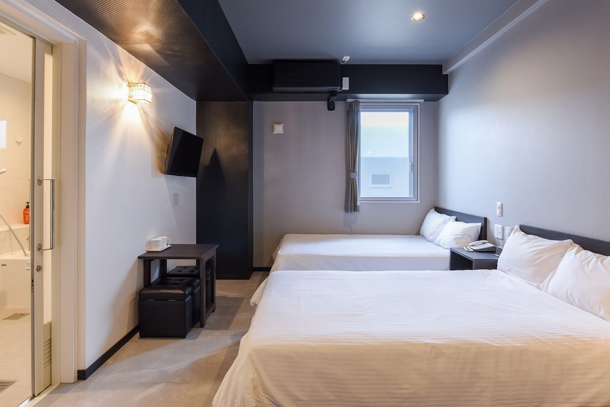 A hotel room with two beds, a TV mounted on the wall, a window with a view, and a small table with a cup and a bottle.