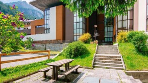 Um edifício moderno com uma escadaria que leva à entrada, cercado por vegetação e uma mesa para piquenique.