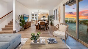 Een ruime woonkamer met een houten salontafel, een bank en een eettafel met stoelen. Links is een trap en aan het einde een groot raam met uitzicht op een zonsondergang.