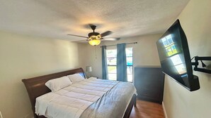 Un dormitorio con cama, ventilador de techo, televisión montada en la pared y ventana con cortinas.