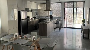Een moderne keuken met een centraal eiland, een eethoek met glazen tafels, en uitzicht op een stenen gevel door grote ramen.