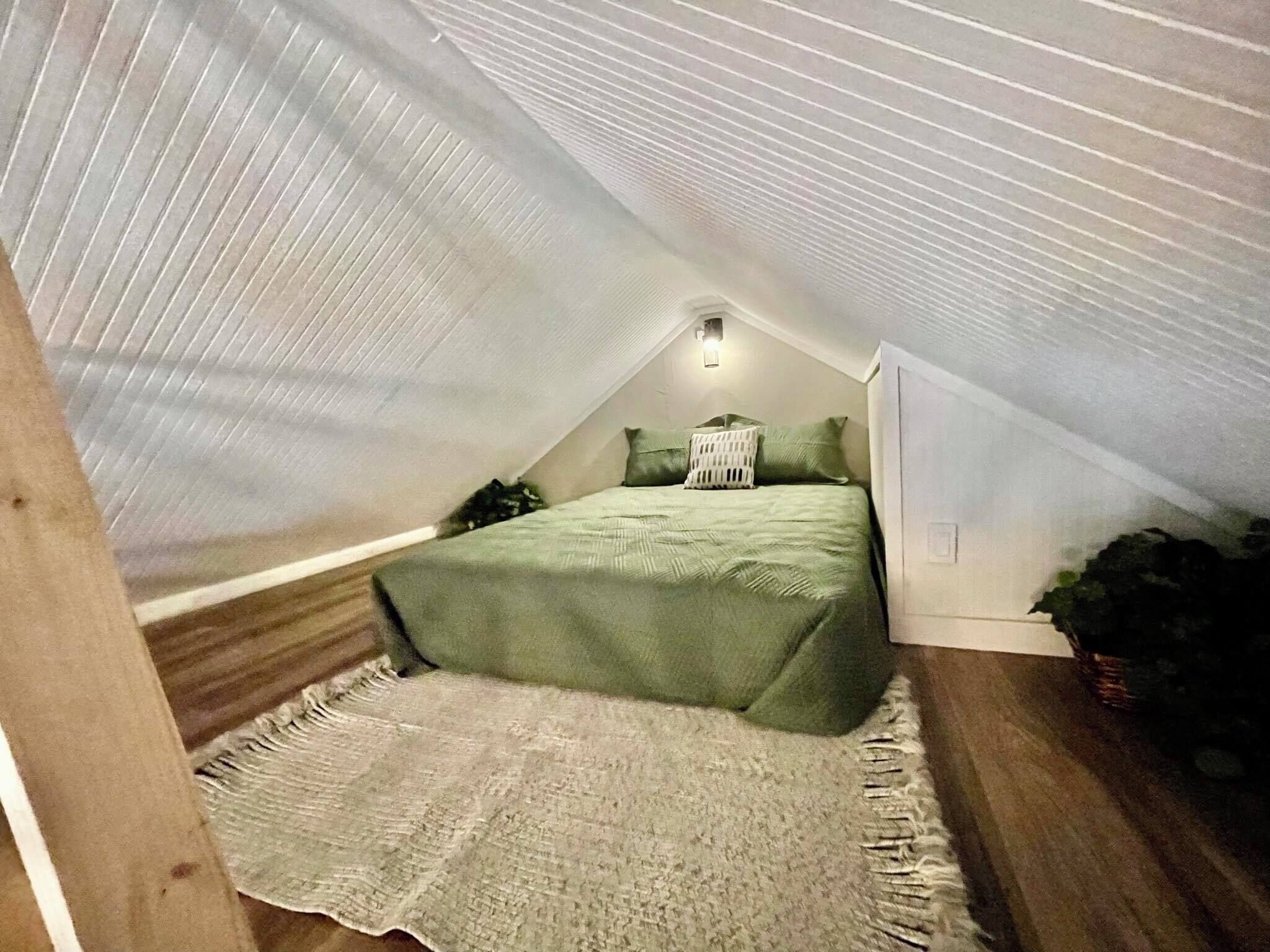 A small, sloped-roofed attic bedroom with a bed, pillows, and a bedside lamp.