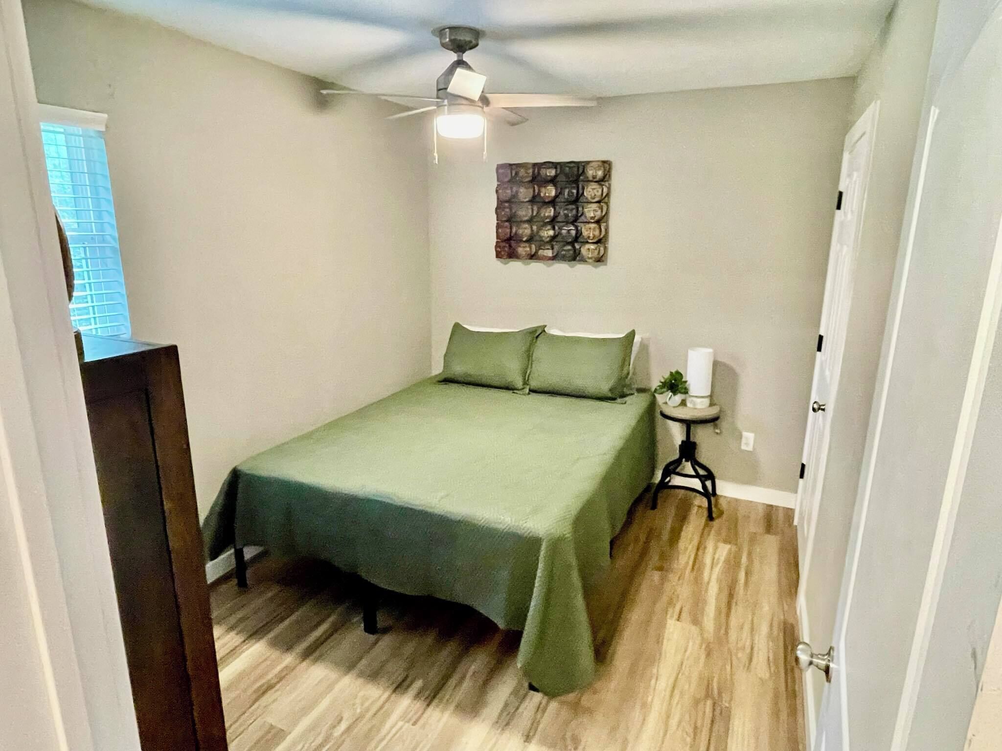 A bedroom with a bed, a ceiling fan, a small table with a plant, and a wall decoration.
