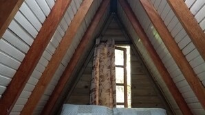 Dormitorio en ático con techos altos, vigas de madera, una ventana con cortina floral y ropa de cama estampada.