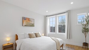 A bedroom with a large bed, a bedside table with a lamp, a window with curtains, and a plant.