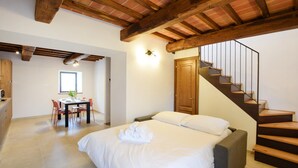 A bedroom with a bed, a wooden staircase, a dining area with a table and chairs, and a view of the outside.