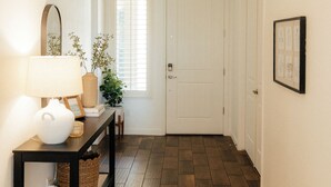 A hallway with a dark wooden floor, a white door at the end, a small table with a lamp and a vase of flowers, and a framed picture on the wall. - Desert Haven W/ Pool, Spa, and Putting Green (Queen Creek)