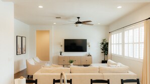 A modern living room with a large flat-screen TV, a ceiling fan, and a dining area with a table and chairs. - Desert Haven W/ Pool, Spa, and Putting Green (Queen Creek)