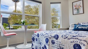 Une chambre à coucher avec un lit, deux chaises, une petite table et une fenêtre donnant sur une tour et des arbres.