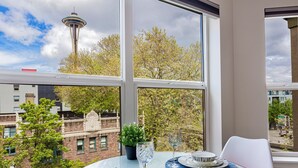 Une table à manger avec des assiettes, des verres et une plante en pot, placée devant une fenêtre offrant une vue sur une tour et des arbres.