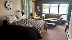 Habitación de hotel con cama, sofá, sillón, ventana con vista a un lago y un reloj en la pared.