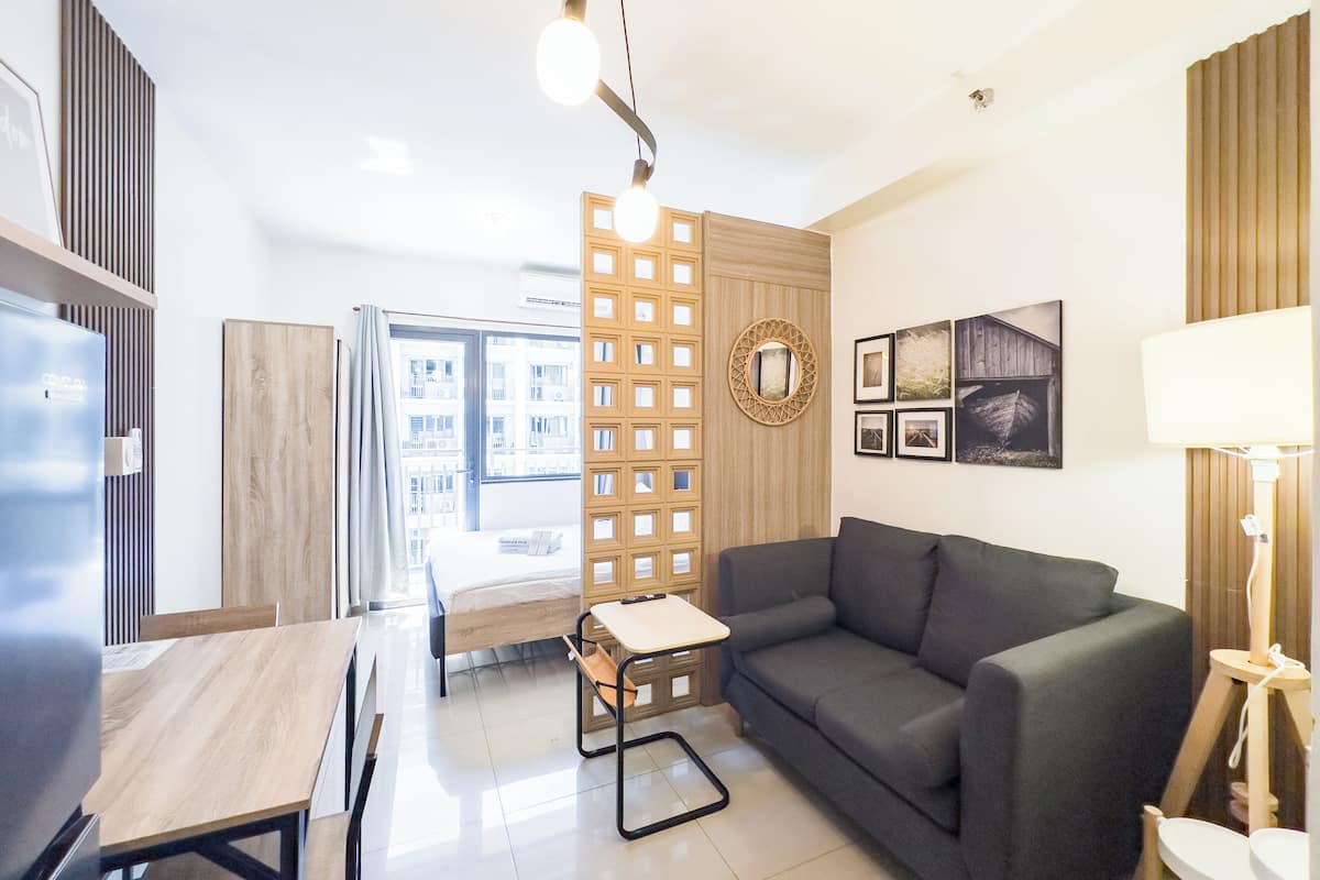 Una sala de estar moderna con un sofá gris, una mesa de comedor de madera y una mesita auxiliar pequeña. Hay una ventana con vistas a edificios, un panel decorativo en la pared y una colección de cuadros enmarcados.