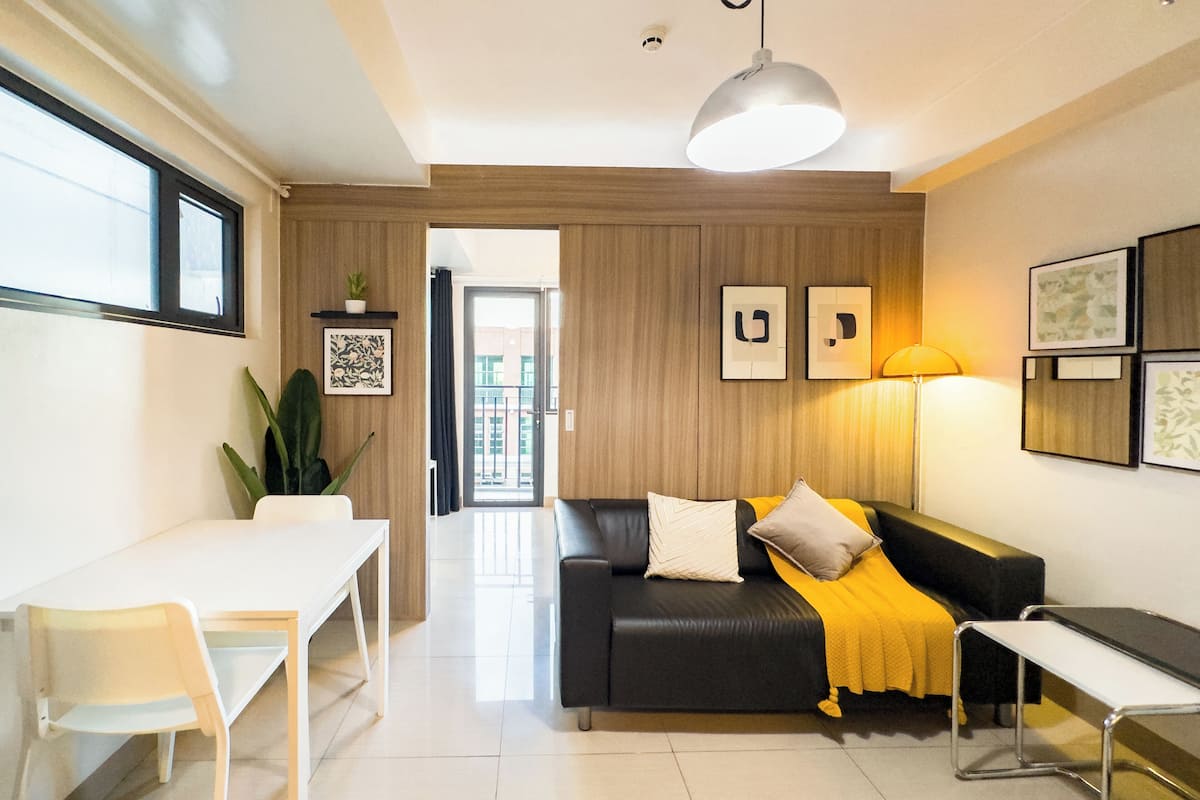 Una sala de estar moderna con un sofá de cuero negro, una mesa blanca y una mesa de centro de vidrio. Se ve un ventanal grande a la izquierda y una puerta que da a un balcón.