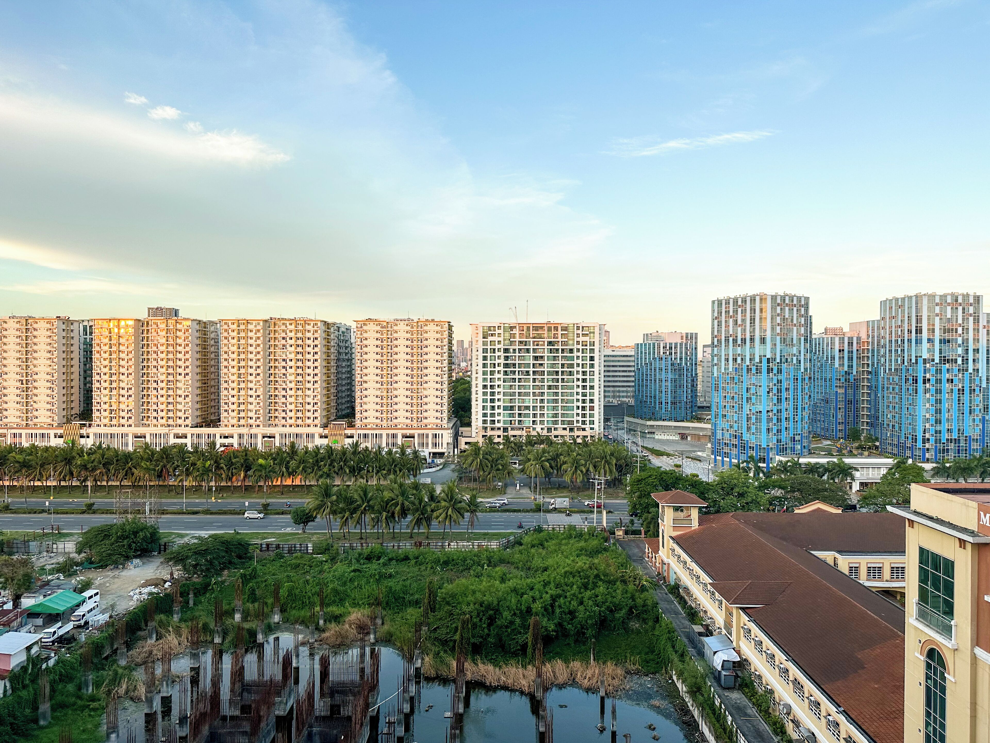 A high-rise residential building complex with multiple apartment buildings, a road with vehicles, and a small body of water with vegetation.