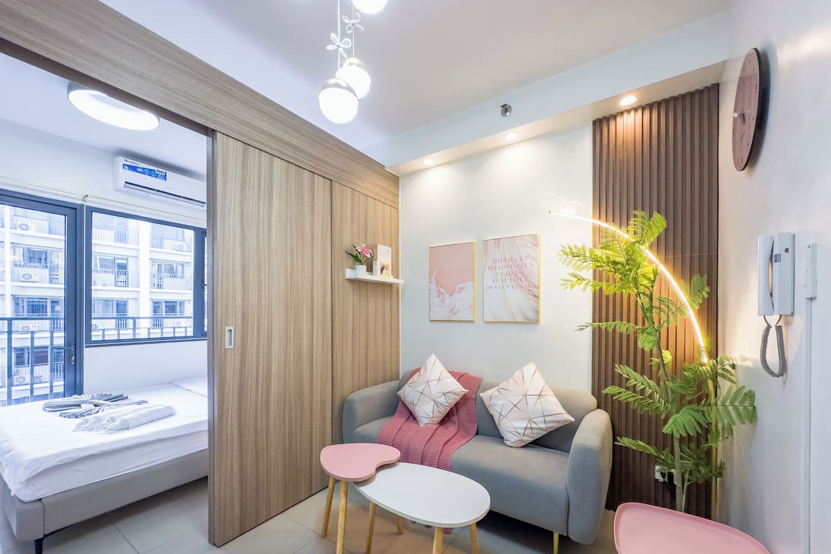 Una habitación de hotel moderna con una cama, un sofá, una mesita y una planta en maceta. Se ve una ventana con vistas a edificios, un teléfono en la pared y una lámpara de techo.