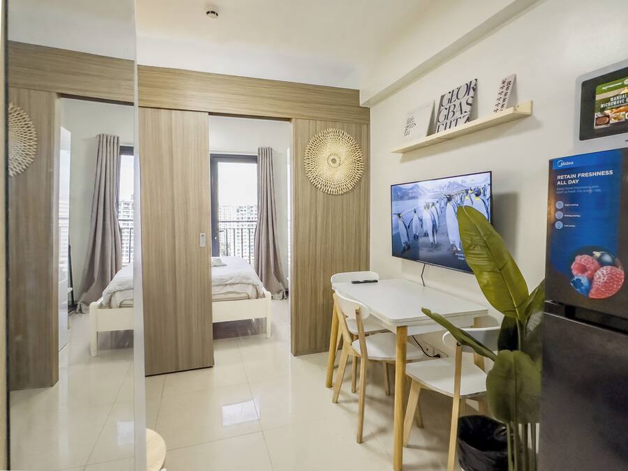 A modern hotel room with a bed, a dining area with a table and chairs, a television, and a plant.