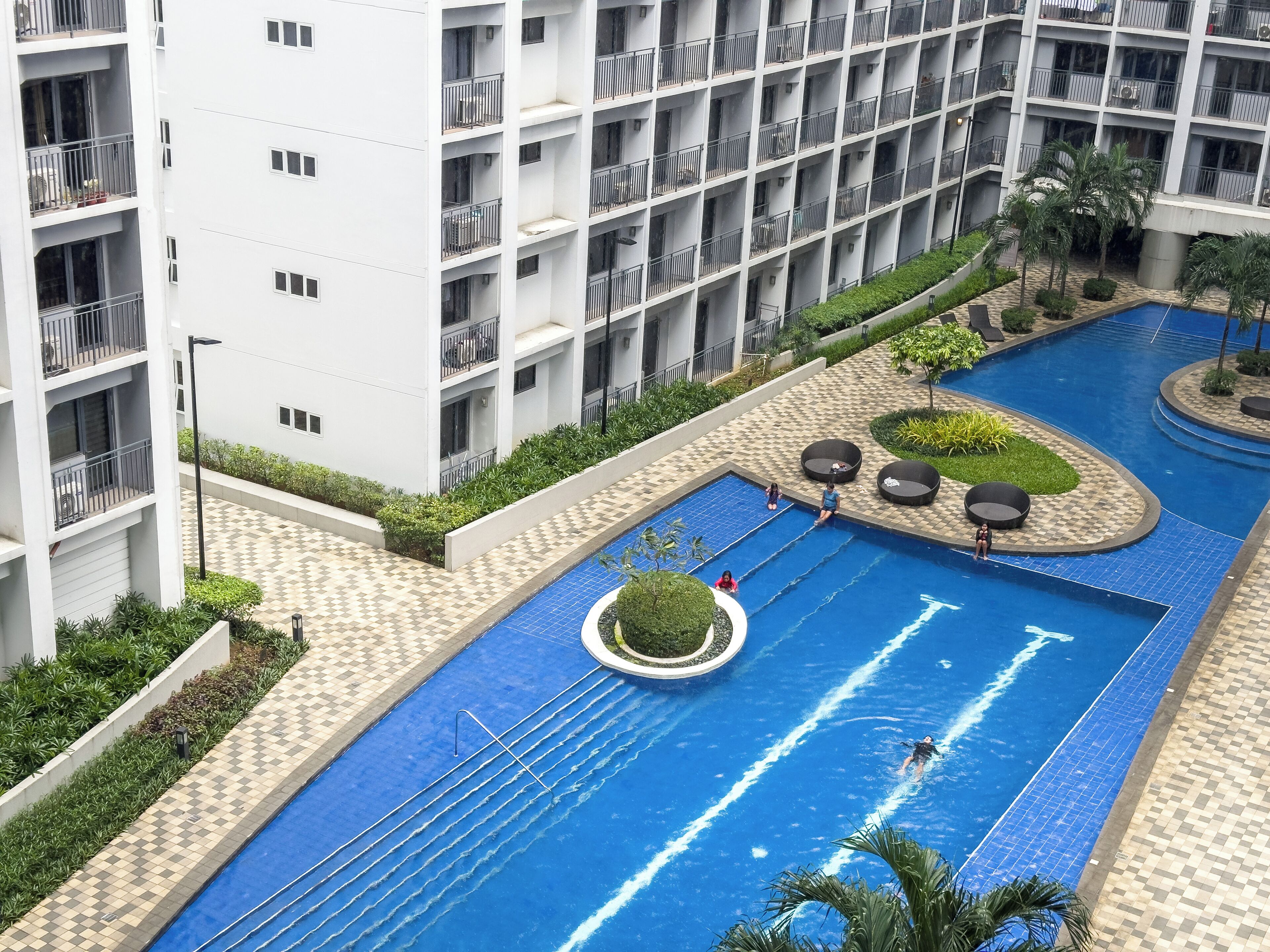 A swimming pool with a tiled deck, surrounded by greenery and a multi-story building with balconies.