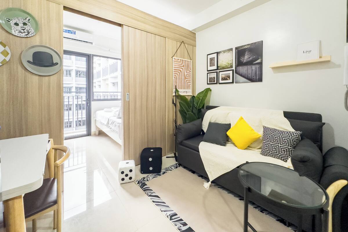 Una sala de estar moderna con un sofá, una mesita y una silla. Se ve una ventana con vistas a edificios.