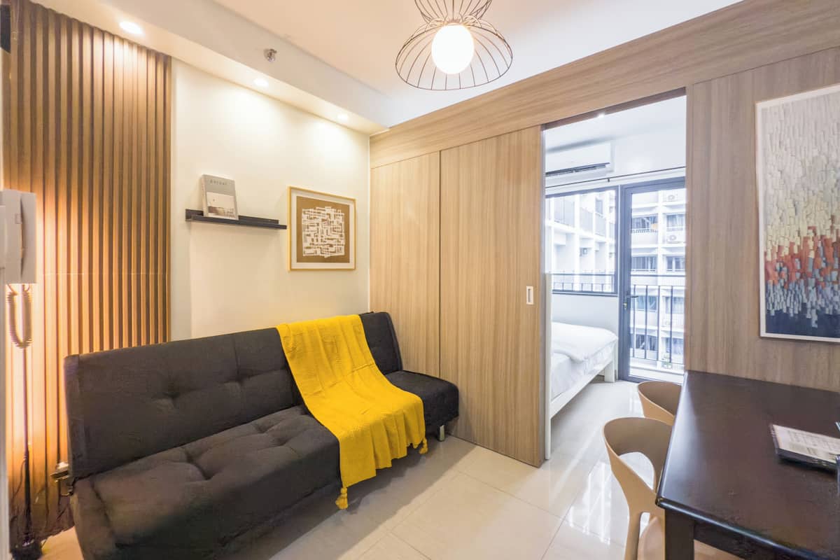 Un salón moderno con un sofá gris oscuro, una manta amarilla, un mueble de madera, una obra de arte abstracta enmarcada y vistas al balcón exterior.