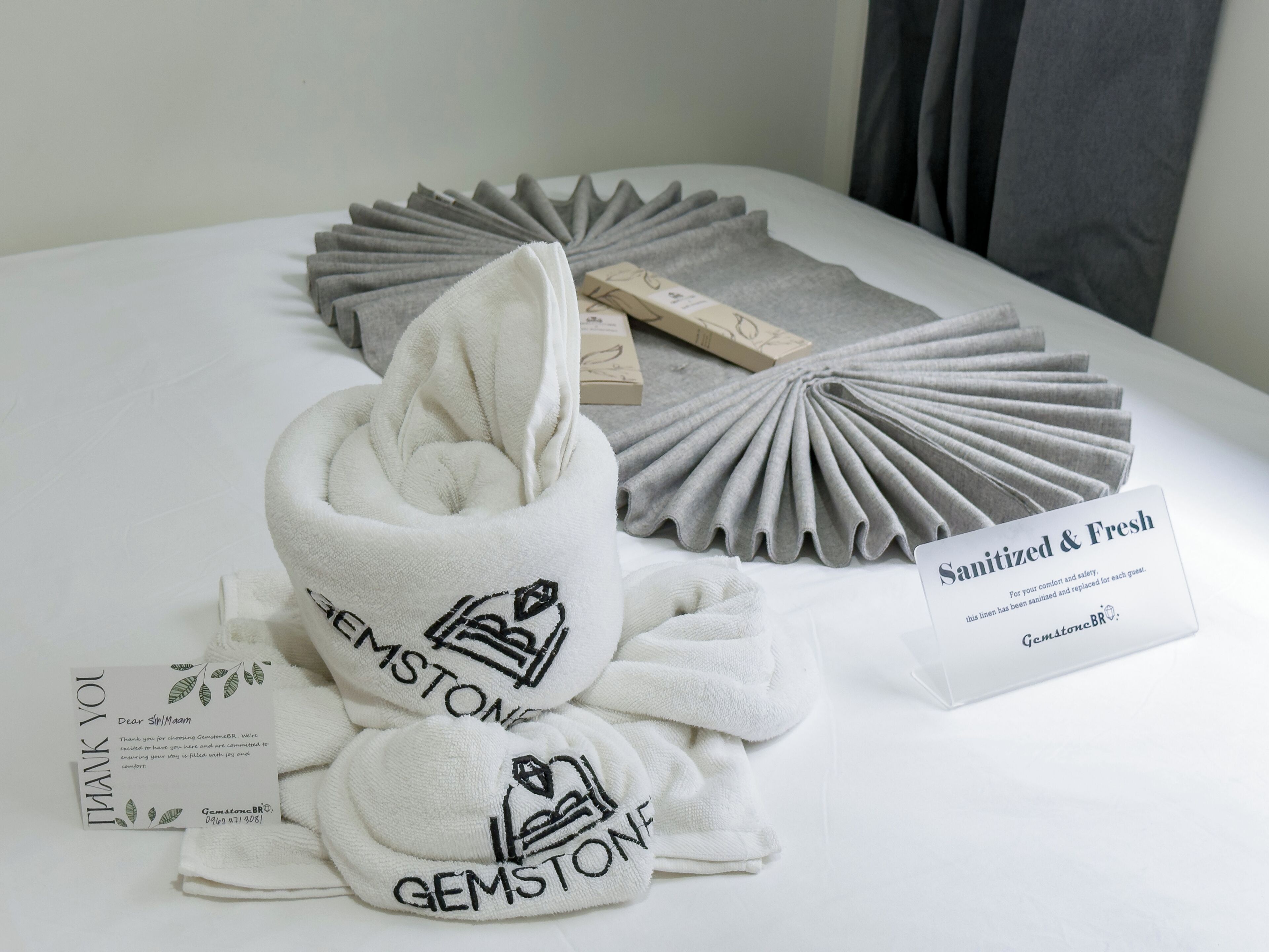 A neatly arranged hotel room with a bed, a folded towel, a tissue box, and a thank you card.