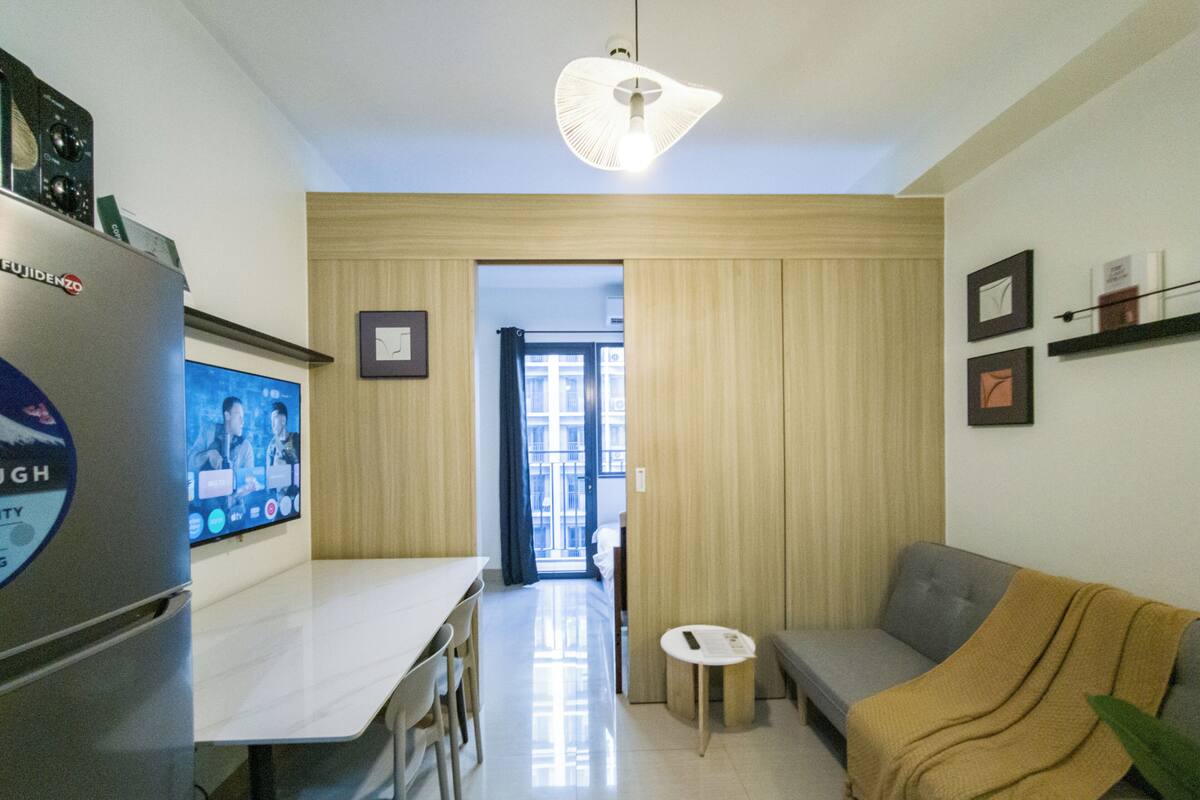 Una cocina compacta con refrigerador, una mesa de comedor con sillas, una mesita redonda pequeña y un sofá con una manta.