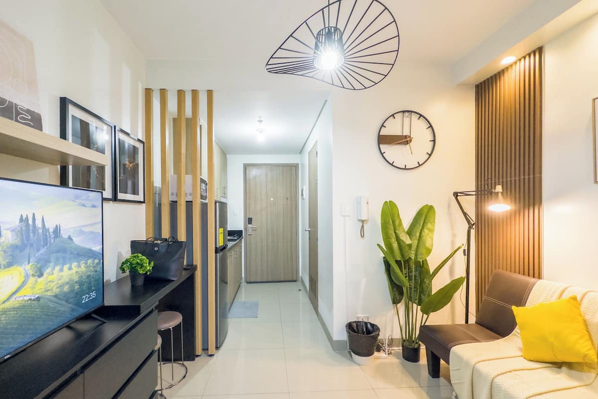 Una sala de estar moderna con un televisor de pantalla plana, un sofá con un cojín amarillo, una planta grande y un reloj en la pared.