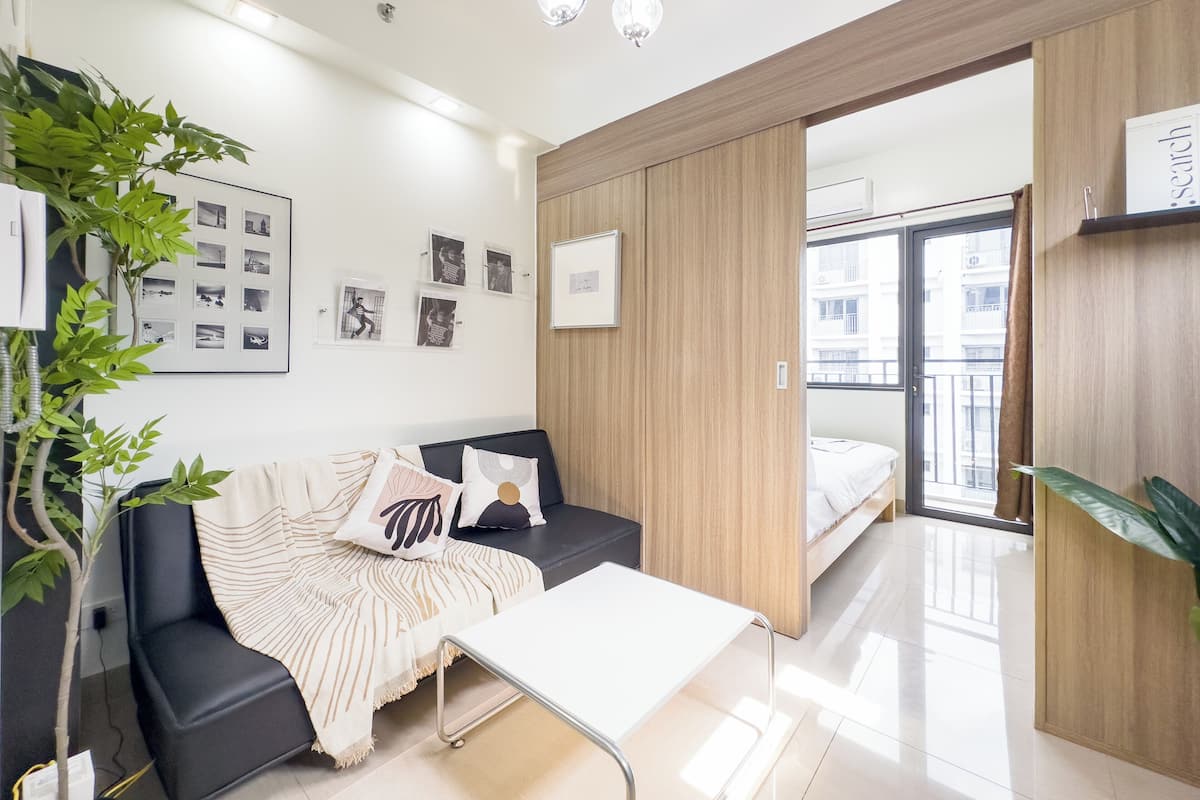 Una habitación moderna y bien iluminada con un sofá negro, una mesa blanca y un panel de madera. Se observan cuadros en la pared y una planta a la izquierda.