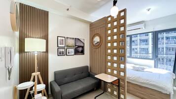 Ein modernes Hotelzimmer mit grauem Sofa, einem kleinen Holztisch, einem Spiegel und einem Bett mit Blick auf die Stadt.