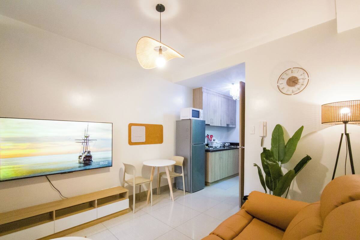 Una sala de estar moderna con una televisión de pantalla plana, un sofá, una mesa de comedor con sillas, una zona de cocina con armarios y un refrigerador, y una planta en maceta.