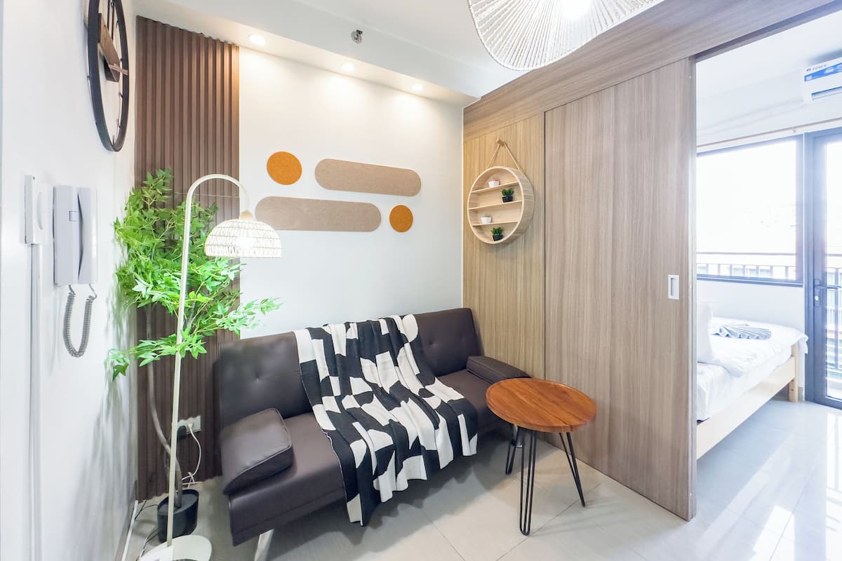 Una sala de estar moderna con un sofá, una mesita redonda, una planta en maceta, un estante de pared con plantas, un reloj y un ventanal grande.