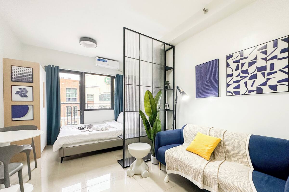 Una habitación de hotel moderna con una cama, un sofá azul con un cojín amarillo, una mesita y una planta en maceta.