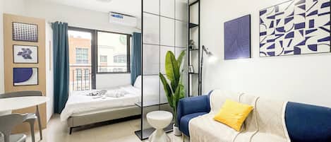 A modern hotel room with a bed, a blue sofa with a yellow pillow, a small table, and a potted plant.