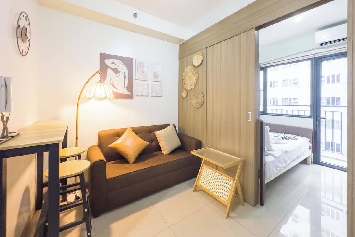 Una habitación moderna y luminosa con sofá, una mesita y una lámpara. Se observan obras de arte en las paredes y una ventana con vistas a edificios.