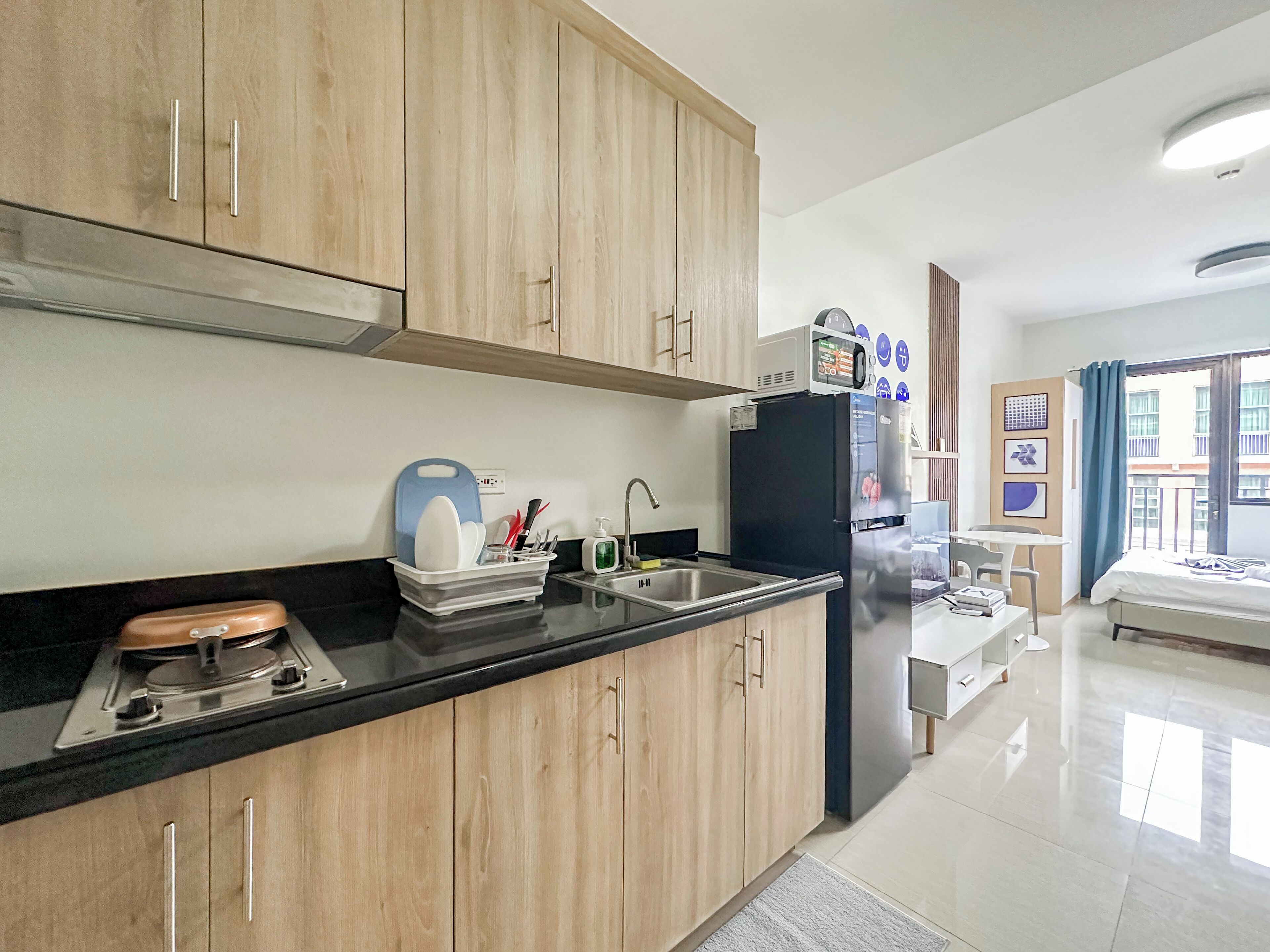 Una cucina moderna con mobili in legno, un piano di lavoro nero e un forno a microonde integrato. Si nota un lavello con rubinetto, un espositore per piatti e un tavolino con una sedia. La stanza ha un'ampia finestra con tende e un letto nell'angolo.